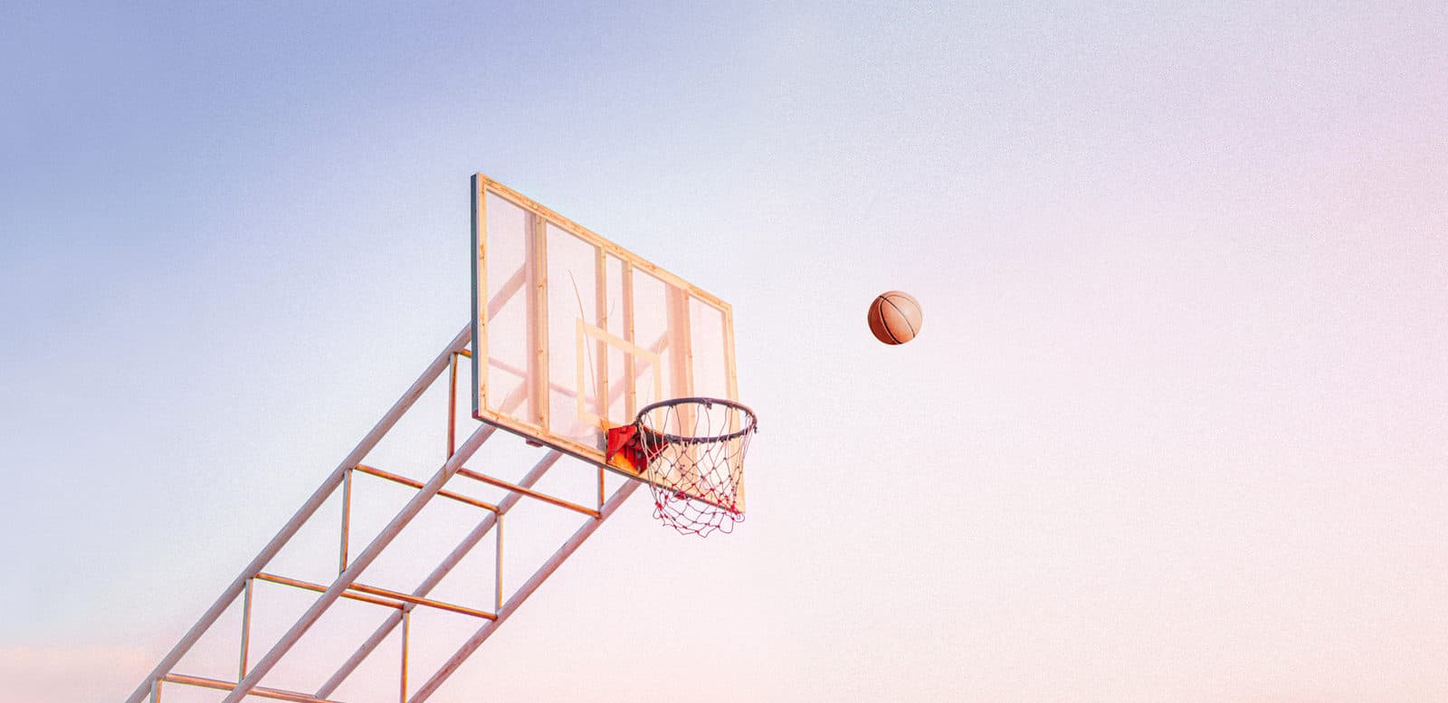 Basketballkurv mot en pastellfarget himmel med en ball på vei mot ringen.