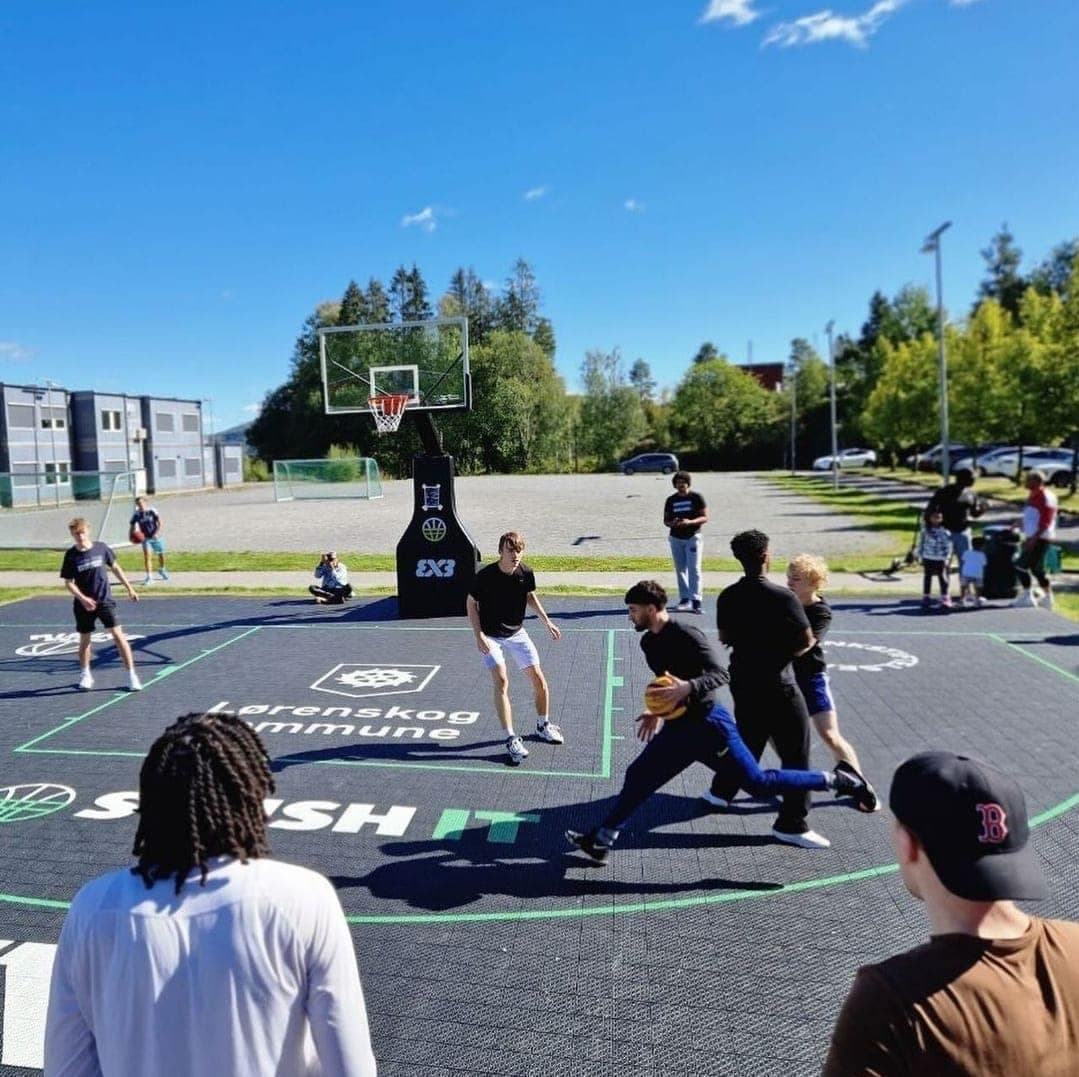 Ungdommer spiller 3x3-basket på en Play IT-bane en solfylt dag, med tilskuere rundt.